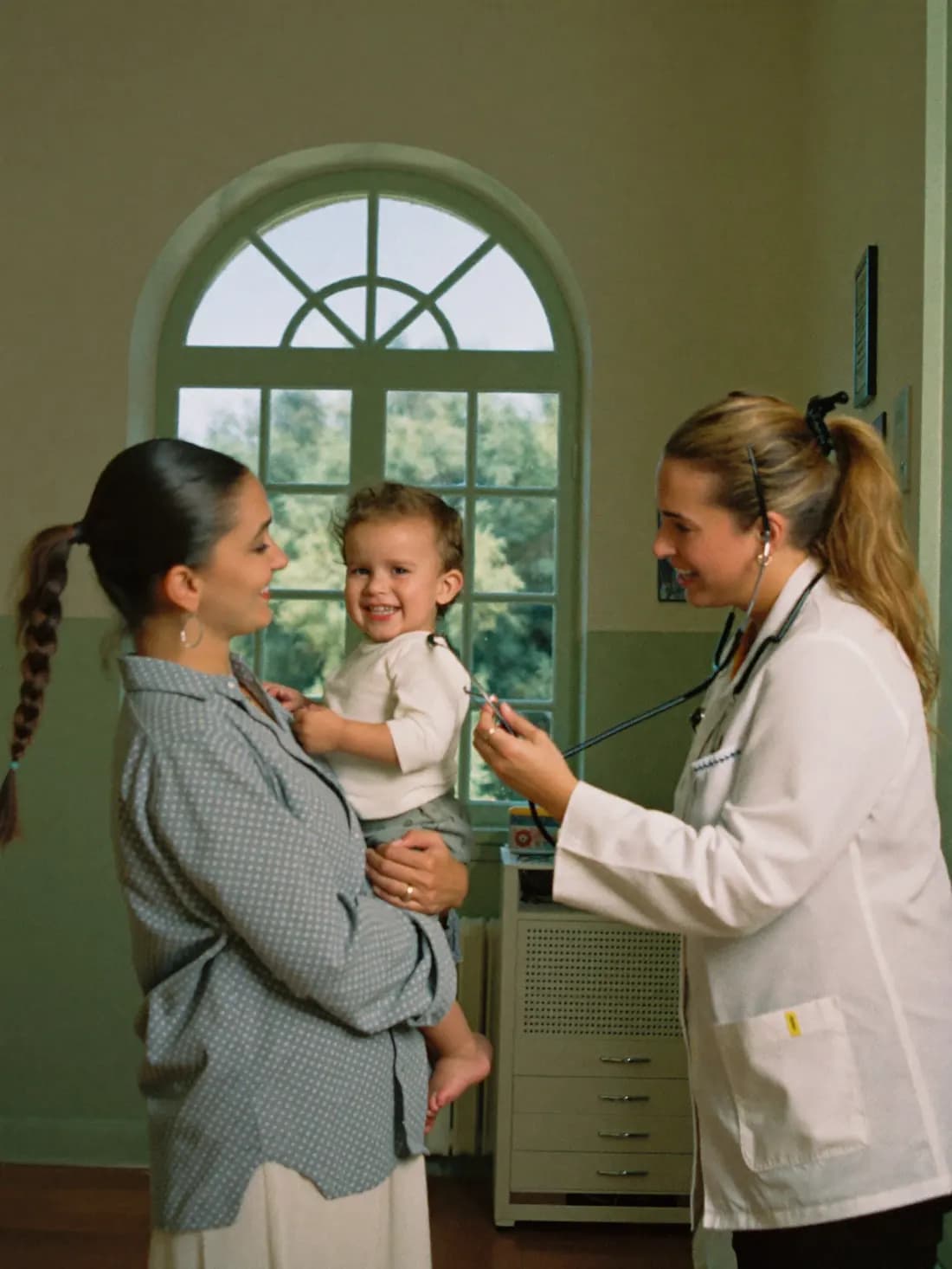 A pediatrician listening to a smiling toddler held by their mother